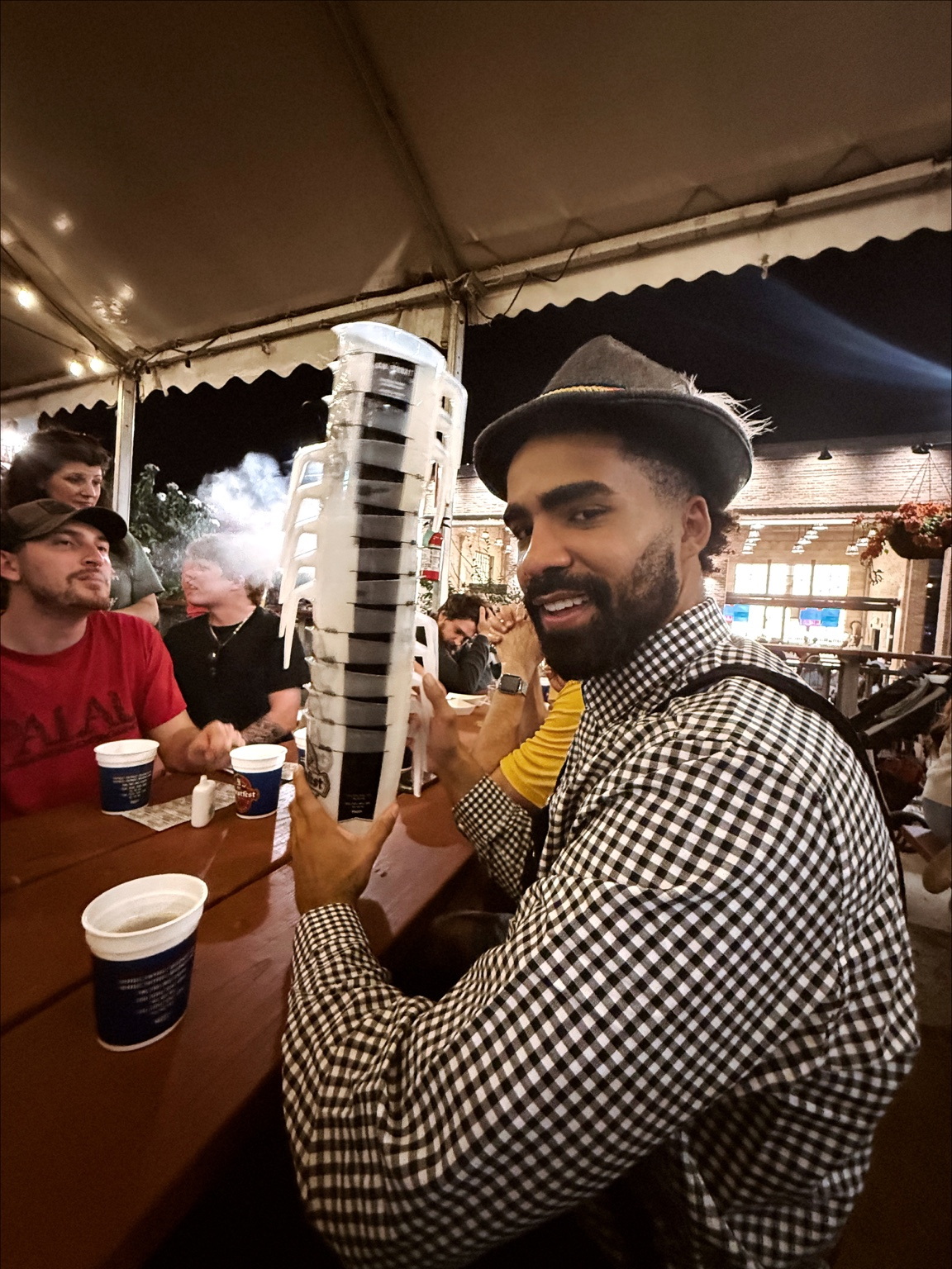 Brandon wearing a checkered shirt and hat at an evening gathering under a tent